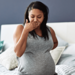 pregnant woman sits on a bed with her hand over her mouth